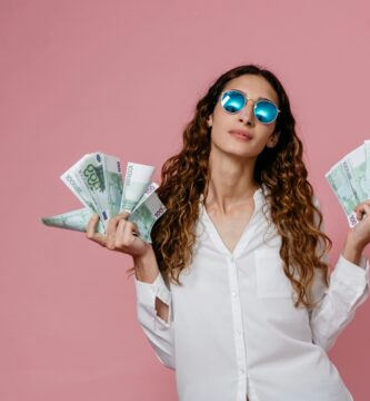 Elegant woman in sunglasses holding euro banknotes against pink background, showcasing confidence and financial empowerment.